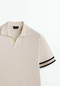 Polo court beige avec col ouvert et manches courtes, avec bordure rayée noire et beige aux poignets.