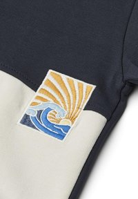 Sweatshirt bleu marine et crème avec un patch brodé représentant des vagues et un soleil en bleu, jaune et blanc. Texture de tissu lisse.