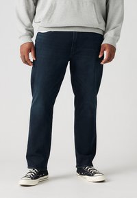 Donkerblauwe spijkerbroek met een rechte pijpdesign, vergezeld van een grijze hoodie. De schoenen zijn zwarte sneakers met witte accenten.