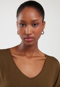 Oliwka zielona bluzka z dekoltem w serek i krótkimi rękawami, gładka w dotyku, z szerokim dekoltem i dopasowanym krojem. Modelka ma na sobie kolczyki w kształcie kół.