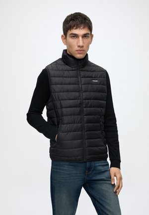 Ung mand iført en sort quiltet vest uden ærmer over en sort langærmet skjorte og blå jeans, står med den ene hånd i vestens lomme.