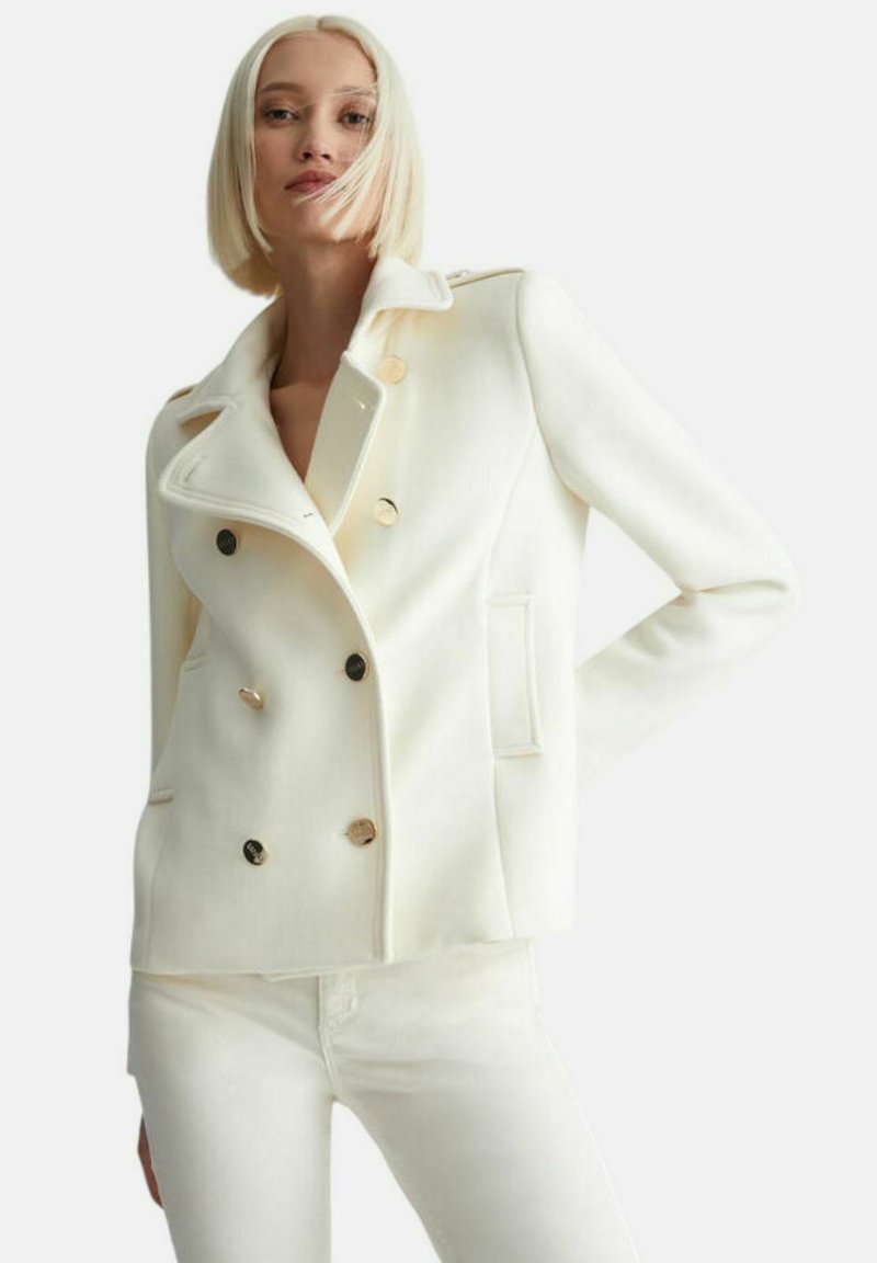 Blazer bianco doppiopetto con bottoni tono oro, rever a forma di scollatura e tasche laterali; realizzato in tessuto morbido. Abbinato a pantaloni bianchi coordinati.