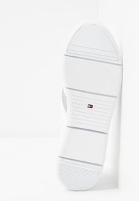 Vit sneaker med en texturerad gummisul, med en silveraccent och en logotyp som säger "TOMMY HILFIGER" på sidan.