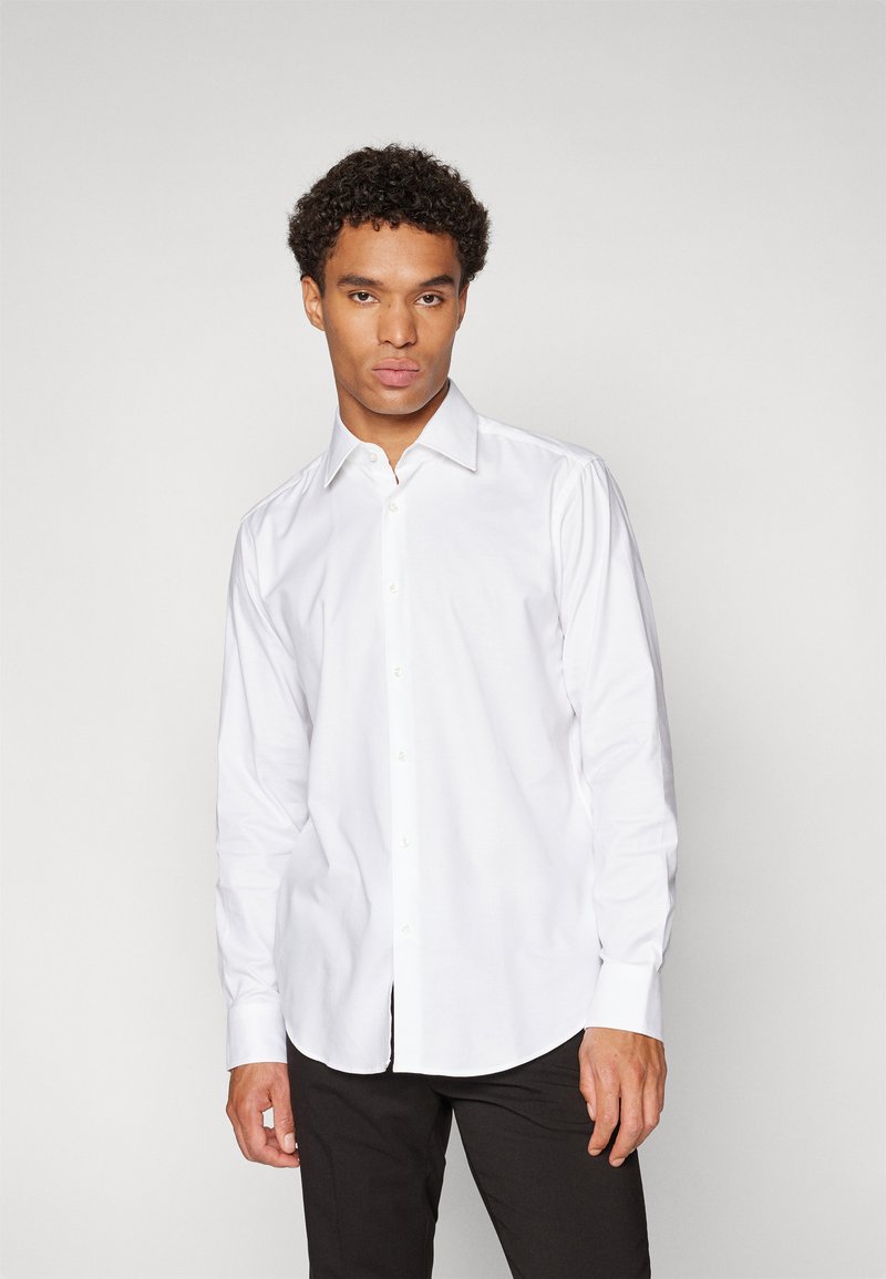 Chemise blanche à manches longues avec col pointu, boutonnière à l'avant et ourlet arrondi ; associée à un pantalon sombre. Texture lisse.