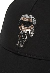 Svart caps laget av stoff med en rhinestone-dekorasjon av en karakter i dress og solbriller på forsiden.