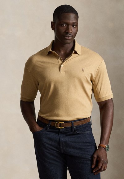 Hombre con camiseta polo beige de manga corta, vaqueros oscuros, cinturón marrón y reloj, de pie con una mano en el bolsillo frente a un fondo neutro.