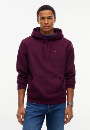 Jeune homme portant un sweat à capuche violet foncé avec une poche devant et un jean bleu, debout devant un fond blanc uni.