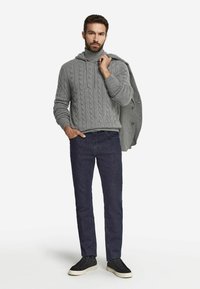 Grauer Strick-Hoodie, dunkle Jeans und schwarze Sneakers. Modell hält einen grauen Mantel und steht vor einem weißen Hintergrund. Einfaches, lässiges Design.