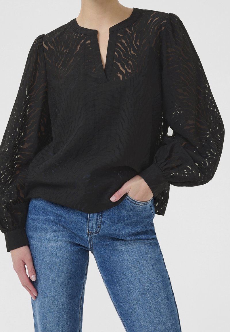 Femme portant un chemisier noir transparent à motifs avec manches bouffantes et un jean en denim bleu, main dans la poche, sur fond clair uni.