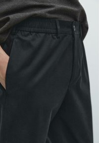 Schwarze Hosen mit elastischem Bund, glatter Textur, seitlichen Taschen und Knopfverschluss. Der Stoff wirkt leicht und elegant.