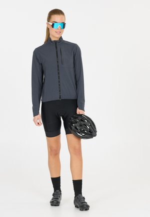 Schwarze leichte Jacke mit hohem Kragen und Front-Reißverschluss, kombiniert mit schwarzen Radshorts. Das Modell hält einen schwarzen Fahrradhelm.