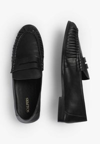 Mocasines de cuero negro con un acabado suave, detalle de costura elevada y tacón plano. Placas y marca visibles en el interior de los zapatos.
