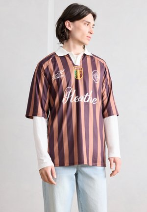 Fiatal férfi barna és narancssárga, függőleges csíkozású, rögbi stílusú ingben, fehér hosszú ujjú felsővel és világos farmernadrágban oldalra fordulva áll.