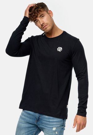 Camiseta de manga larga negra con cuello redondo, material de algodón suave y un logotipo blanco en el pecho. Llevada con jeans vaqueros claros desgastados.