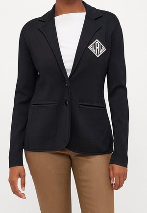 Sort skræddersyet blazer med to knapper, stiklommer, en hvid indertrøje og beige bukser på en stående person.