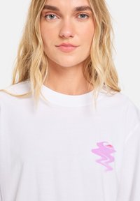 Weißes Baumwoll-T-Shirt mit Rundhalsausschnitt, das ein kleines pinkes abstraktes Design auf der linken Brustseite aufweist. Weicher Stoff und lockere Passform.