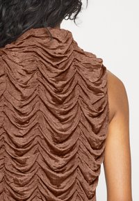 Brun texturerad plagg med lager av plisseringar som skapar ett vågformat mönster. Tyget verkar mjukt med en lätt glans.