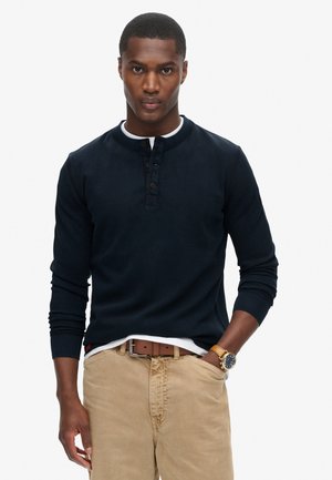 Man in een marineblauw henley-shirt met lange mouwen, beige broek met een bruine riem en een horloge met gouden kleur, staand tegen een effen witte achtergrond.