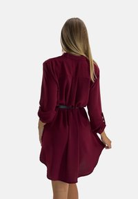 Robe chemise bordeaux à manches longues, taille ceinturée et jupe fluide. Fabriquée en tissu léger et texturé avec des accents de boutons sur les manches.