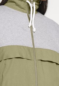 Chaqueta de cremallera verde y gris con un alto cuello, que presenta un cordón ajustable, cierre completo de cremallera y un diseño de tela en dos tonos.