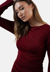 Burgundowa dopasowana bluzka z długim rękawem i okrągłym dekoltem, z marszczonymi detalami po bokach, które podkreślają smukłą sylwetkę.