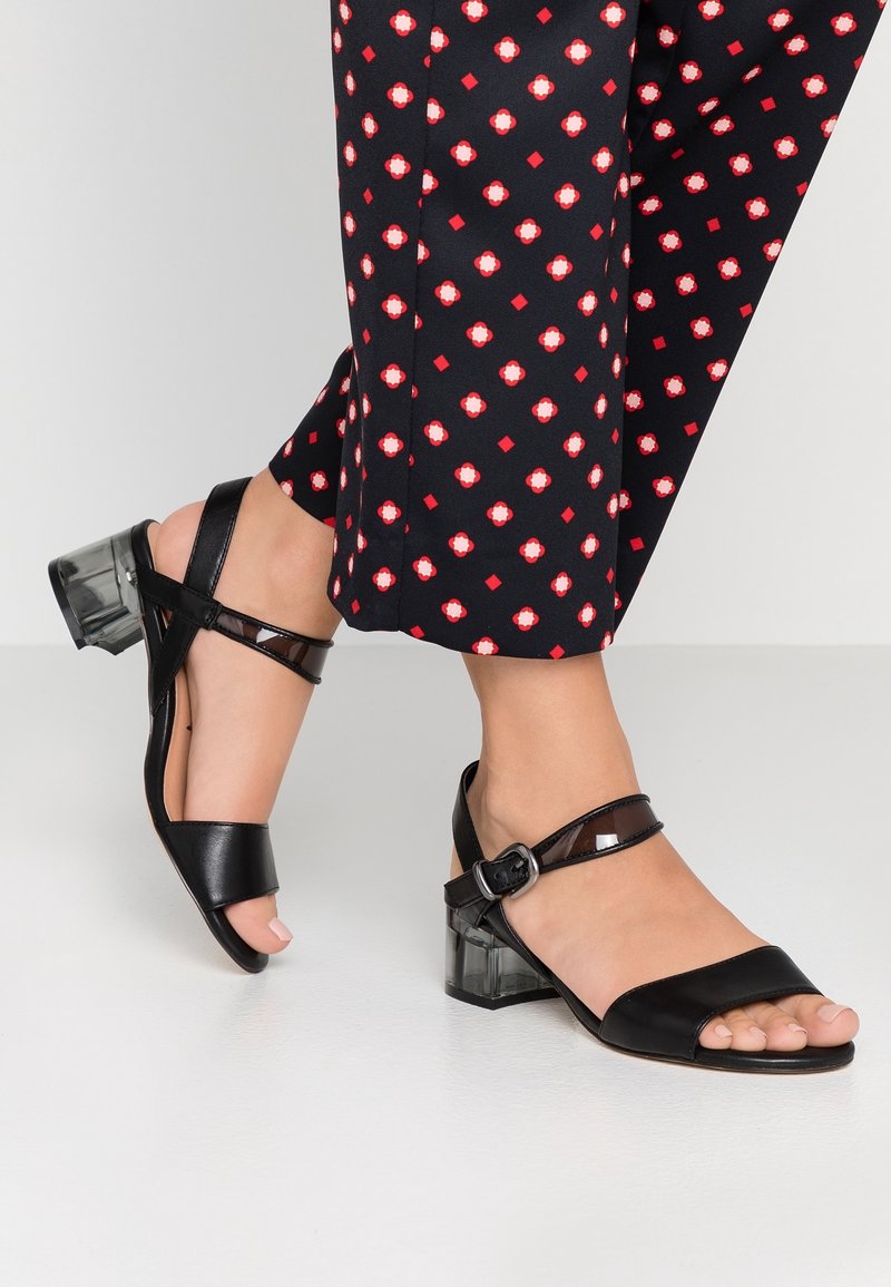 Sandales noires à bout ouvert avec une large sangle couvrant le cou-de-pied et une sangle de cheville ajustable. Dotées d'un talon bloc transparent.