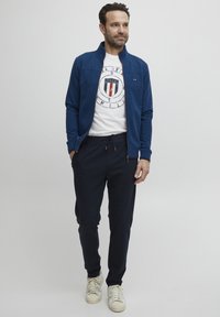 Marineblå zip-up jakke over en hvit t-skjorte med grafikk, kombinert med marineblå joggebukser og kremfargede joggesko; har logo-detaljer og snøring i livet.