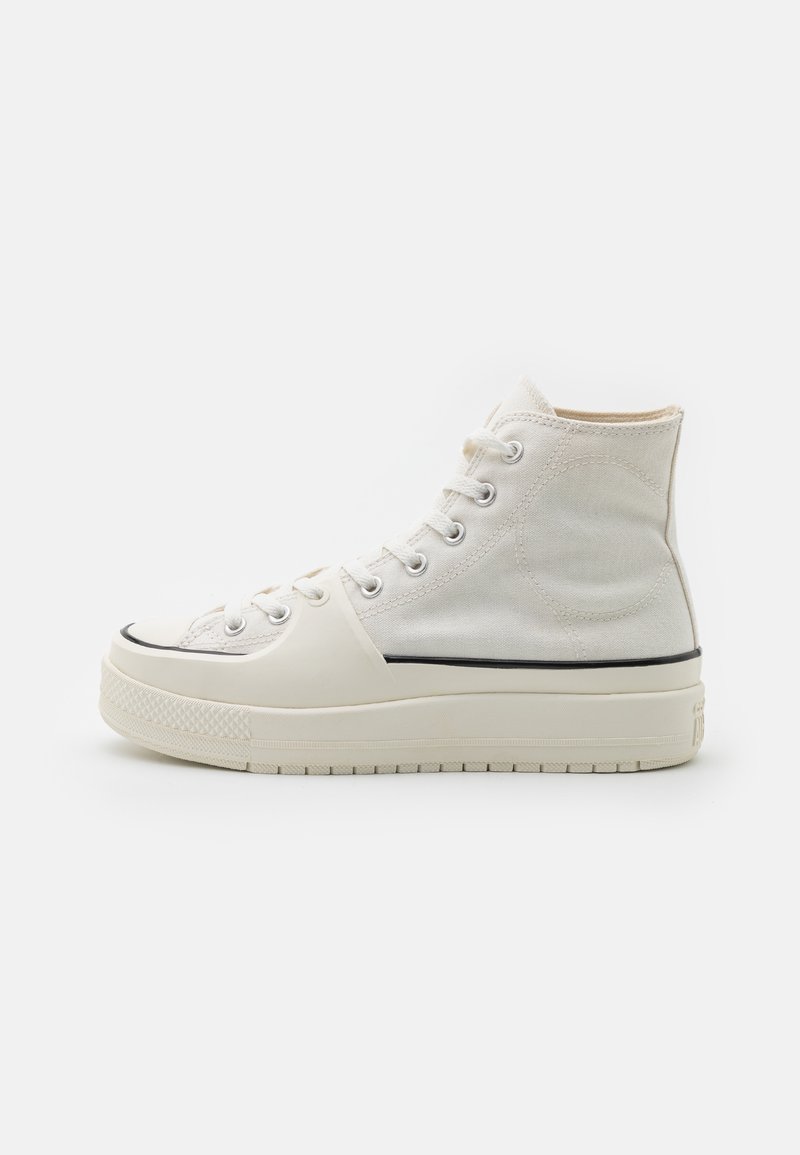 High-top sneakers in off-white canvas with a rubber sole, featuring black trim and metal eyelets, with textured fabric on the upper.