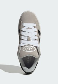 Grauer Wildledersneaker mit weißen Schnürsenkeln, schwarzen Akzenten und einem auffälligen Logo auf der Zunge, mit strukturierter Oberfläche und gepolstertem Innenfutter.