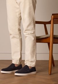 Marineblauwe suede instappers met witte rubberen zolen, gecombineerd met crèmekleurige broeken, staand op een houten vloer naast een houten stoel.