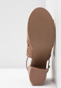 Chaussure à talon haut en suède marron vue de la semelle, reposant verticalement contre un mur blanc et un sol blanc.