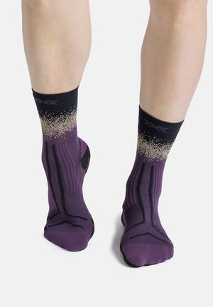 Een paar benen met paarse en zwarte crew-sokken met een beige gespikkeld patroon vlak bij de bovenkant, staand op een witte achtergrond.
