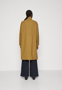 Cappotto lungo e leggero di colore giallo senape, con colletto alto, vita elastica e tasche laterali. Abbinato a jeans scuri a gamba larga.