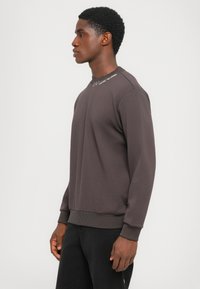 Mørk grå sweatshirt med rund hals, ribbede mansjetter og hem. Har subtil merking på halsen. Stoffet ser mykt og teksturert ut.