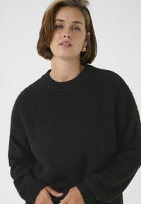 Schwarzer, strukturierten Pullover mit rundem Ausschnitt und lockerer Passform. Der Stoff wirkt weich und gestrickt, mit langen Ärmeln und gerippten Bündchen.