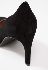 Escarpin en daim noir à talons hauts avec un talon pointu et un dos fermé sur fond blanc.