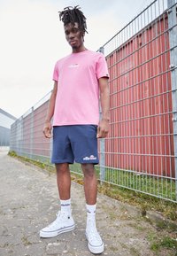 Rosa Baumwoll-T-Shirt, marineblaue Shorts mit Logo, weiße High-Top-Sneaker und weiße Socken. Außenbereich mit einem Zaun und rotem Hintergrund.