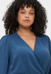 Top bleu enroulé mettant en avant un décolleté en V avec une bordure en dentelle festonnée, des manches courtes bouffantes et un tissu texturé. Matériau lisse et léger.