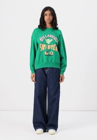 Felpa verde in cotone con stampa di testo e grafiche, abbinata a pantaloni larghi in denim blu scuro e sneaker bianche.