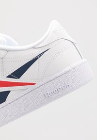 Vit Reebok-sneaker med texturerad sula, marinblå och röda sidostripes, och präglad Reebok-logotyp på hälen och sulan.