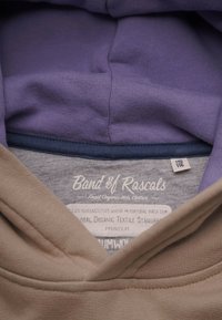 Vaalean lilac ja beige huppari, jossa on pehmeä rakenne. Tunnusalaalla on teksti, joka ilmoittaa brändin, tuotetiedot ja orgaanisen sertifioinnin.