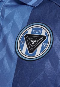 Zila sporta krekls ar dimantu rakstu, apaļa uzlīme ar trīsstūra dizainu un tekstu "XXIII" kontrastējošā krāsā.