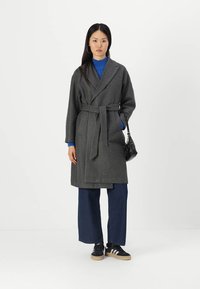 Grå ullblanding omslagsfrakk med belte, skjortekrage og sidelommer, brukt over en blå ribbestrikket genser og vide denimbukser.