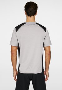 Grå sportt-shirt med svarta detaljer på axlarna, korta ärmar och rundad nederkant. Har logotypen "J. Lindeberg" på övre delen av ryggen.