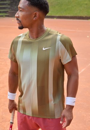 Olive green athletic shirt with a geometric pattern, short sleeves, and a Nike logo, paired with pink shorts and a tennis racquet.