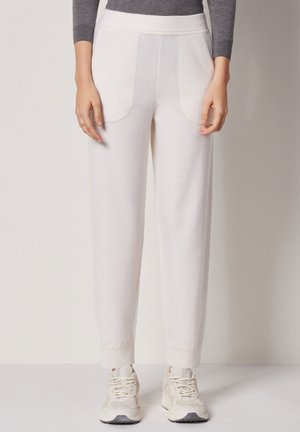 Person iført hvide strikkede bukser med forlommer, grå langærmet top og hvide snørebåndssneakers, stående mod en ensfarvet baggrund.