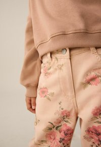 Világos rózsaszín virágos nadrág finom rózsamintákkal és zöld levelekkel, bézs pulóverrel párosítva. Simogató textúra, hétköznapi kialakítás.