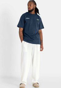 Camiseta de algodón azul marino de mangas cortas, con acentos de texto en blanco. Combinada con pantalones blancos de corte holgado y calzado beige.