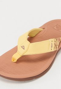 Sandale flip-flop galbene cu talpă din cauciuc, tălpicul texturat de culoare maronie și curea cu model ce prezintă un logo. Design ușor și casual.