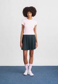 Różowa dopasowana koszulka, ciemnozielona plisowana spódnica, różowe skarpetki z czarnym logo i białe buty. Prosty, sportowy design z miękkimi teksturami.